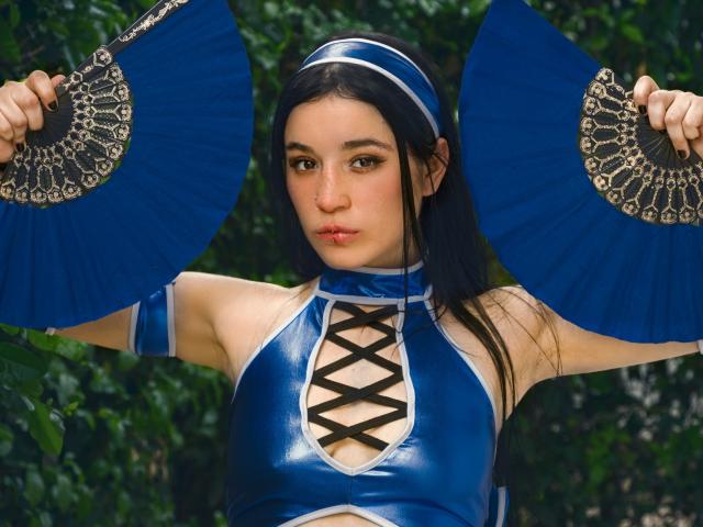 DulceHarper con un abito di lattice blu, un corpetto nero con lacci, che tiene ventagli blu con motivi elaborati, lunghi capelli neri, una fascia per capelli, uno sfondo con foglie verdi, un'espressione accigliata, un trucco leggero, un aspetto sensuale..