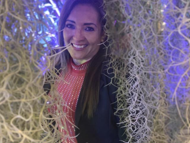 FreyjaCam with long brown hair, smiling, wearing a red dress with white polka dots, black jacket, surrounded by tangled, golden vines, in a purple-lit environment.