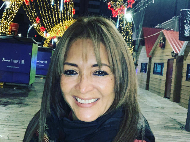 FreyjaCam with long brown hair, smiling, wearing a black scarf, showing her white teeth, and blue eyeliner, standing on a wooden boardwalk at night with festive lights.