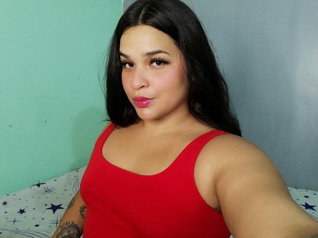 Antonela, com cabelos pretos longos, blusa de alça vermelha, batom rosa, tatuagem no braço direito, parede verde clara e cinza, tirando uma selfie, mostrando os seios e o rosto bonito..
