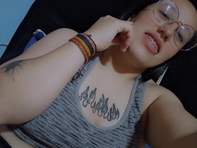 NatalieHarris with glasses, tongue out, gray ribbed tank top, "I am" tattoo on chest, colorful bracelets, black hair, tattoo on left arm, smiling, light skin.