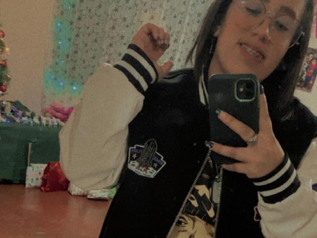 Natalie Harris, de óculos, sorrindo, vestindo uma jaqueta esportiva preta e branca, tirando uma selfie com um telefone verde, braço direito levantado, mostrando uma expressão fofa e confiante..