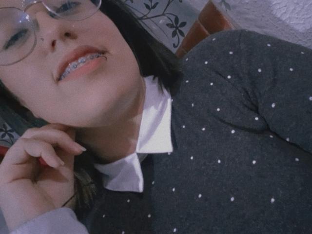 NatalieHarris with glasses, dark hair, grey polka-dot shirt, white collar, smiling with metal braces, holding her chin, soft light, close-up, casual, slightly blurry.