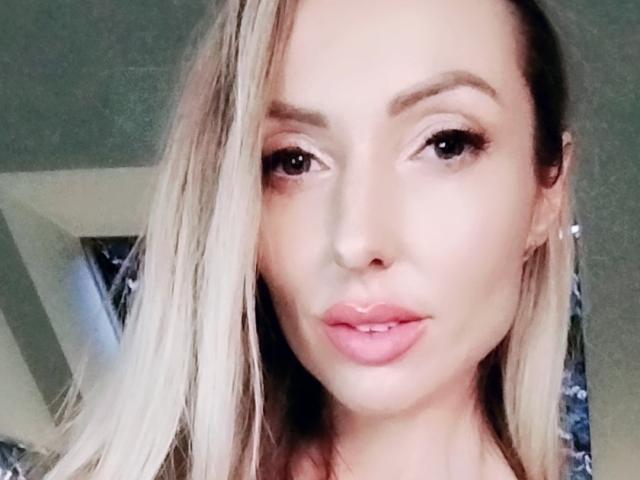 SexyEmila heeft blond haar, volle lippen met roze lippenstift en bruine ogen. Ze kijkt naar de camera met een lichte glimlach, haar gezicht is gecentreerd en in close-up..