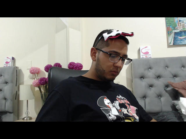 Erico Jones, con cabello negro corto, gafas, camiseta negra con Mickey Mouse, diadema roja y blanca con forma de estrella, sentado en una silla gris con capitonado, mirando hacia abajo, la mano derecha fuera del encuadre..