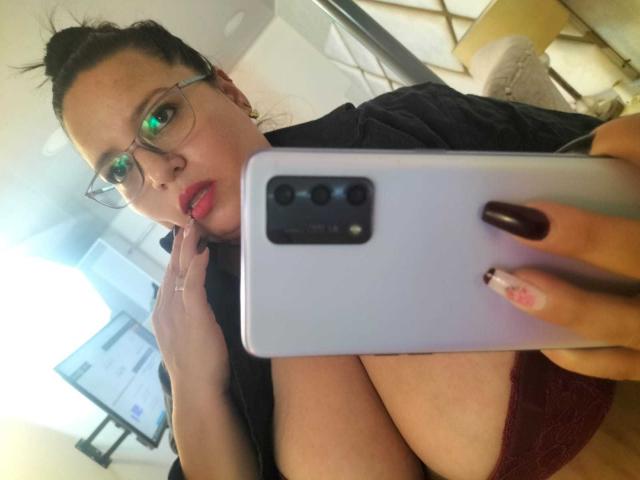 SkayLaraWalker, avec des lunettes, du rouge à lèvres et des ongles foncés, tenant un téléphone devant ses seins, portant une chemise noire et un soutien-gorge rouge, dans un autoportrait..