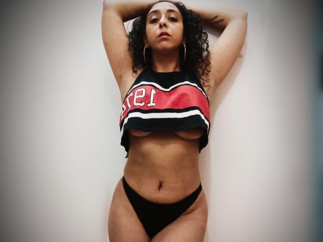 KellyLoveOF met krullend haar, draagt een rode en zwarte crop top die haar borsten laat zien en zwarte onderbroek, met armen omhoog, staand tegen een witte muur. Zeer zelfverzekerd, sexy..