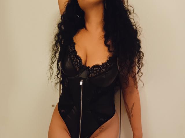 KellyLoveOF met krullend zwart haar, gekleed in een zwart, doorschijnend bodypak met een ritssluiting aan de voorkant, waardoor haar borsten zichtbaar zijn en een tatoeage op haar rechter dijbeen te zien is, voor een witte muur..
