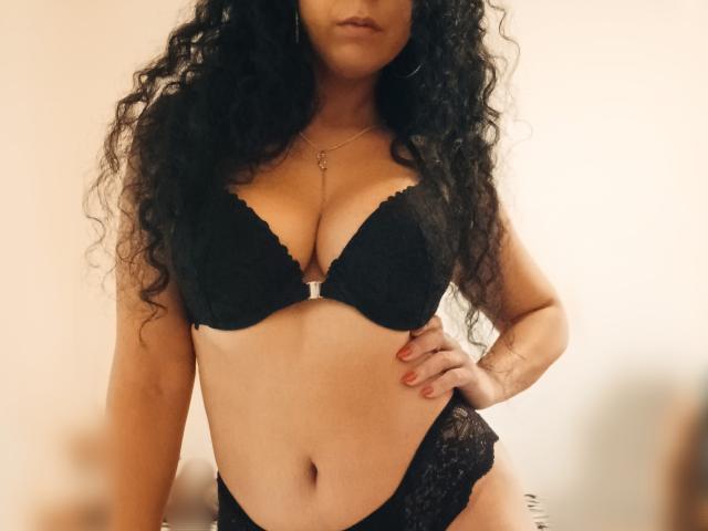 KellyLoveOF met krullend zwart haar, draagt een zwarte kanten bh en bijpassende onderbroek, toont haar borsten en haar geslachtsdeel bedekt met kant, hand op haar heup, ketting, rode nagels..