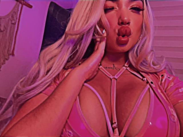 YaraPolly with platinum blonde hair, pouty lips, and pink strappy lingerie, showing off her large tits, hand near face, pink lighting, and a tattoo on her chest.