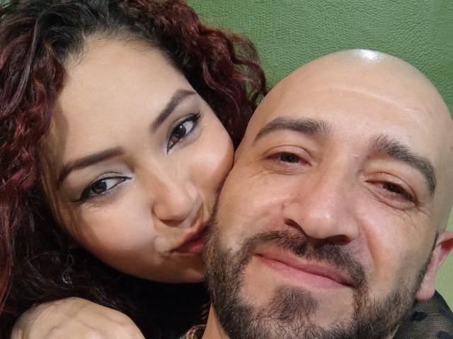 Catelyn tiene el pelo rizado y pelirrojo, delineador de ojos negro, y está frunciendo los labios cerca de la cabeza calva de Fer, que tiene barba. Fer está sonriendo con una ligera sonrisa..