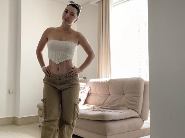 Valery Lombardi, con las manos en las caderas, vistiendo un top blanco sin tirantes que deja ver sus pechos y pantalones cargo color beige, de pie frente a un sofá beige, sonriendo..