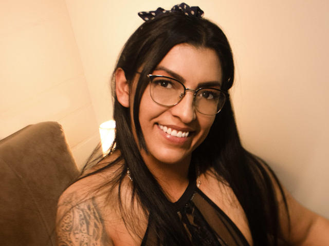 JanetteRider, avec ses longs cheveux noirs, ses lunettes et son haut en dentelle noire, sourit à l'appareil photo, montrant un tatouage sur son bras droit, et porte un élastique pour les cheveux noir..