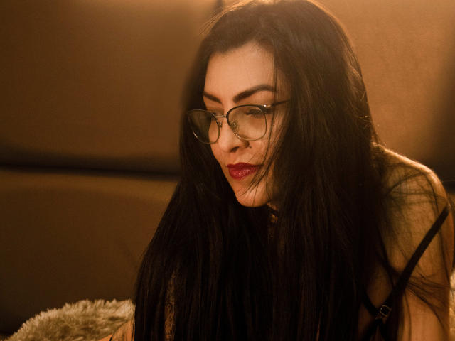 JanetteRider, avec ses longs cheveux noirs, ses lunettes et son rouge à lèvres rouge, regarde vers le bas avec une expression sérieuse, portant un haut noir à bretelles, dans un environnement chaleureux, éclairé en brun..