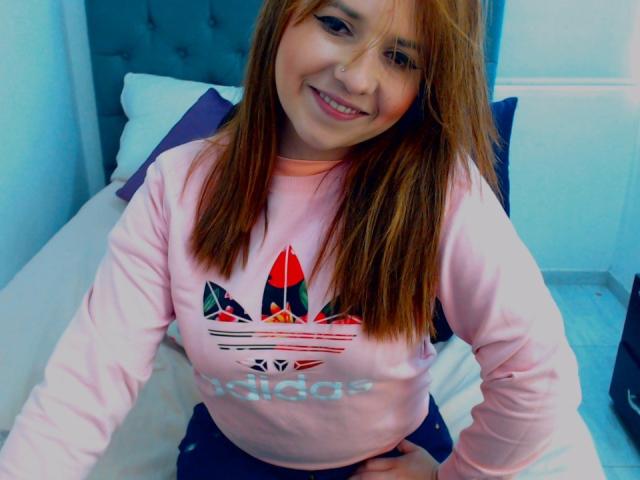 Kkatalina with light brown hair, smiling, wearing a pink Adidas shirt with a red and black logo, sitting on a bed, hands on her knees, tits slightly visible.