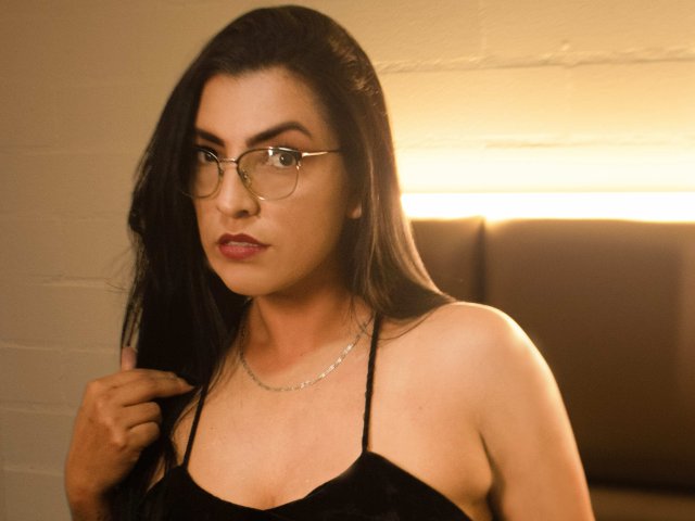 JanetteRider, avec ses cheveux noirs, ses lunettes et son rouge à lèvres rouge, portant un haut noir à bretelles fines, touchant ses cheveux, mettant en valeur sa poitrine et un collier en argent, dans une lumière chaude..