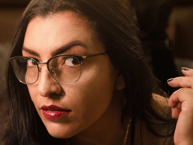 JanetteRider with dark brown hair, wearing glasses, red lipstick, and black nail polish, holding her hair with her right hand, looking slightly to the side.