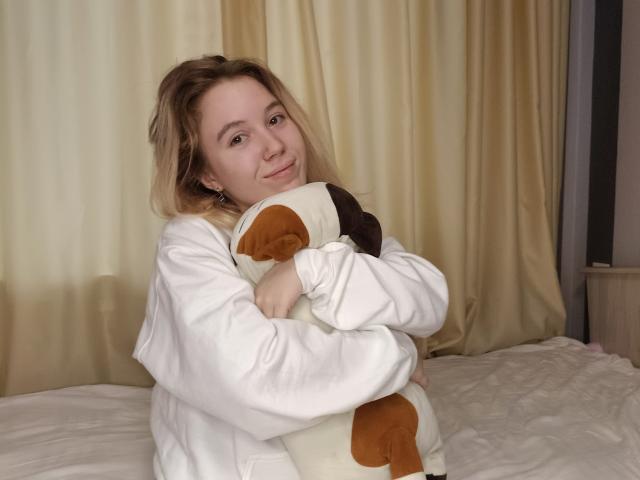 Anika, avec des cheveux blonds, une chemise blanche, tenant un chien en peluche marron et blanc, souriant doucement, assise sur un lit avec des rideaux beige en arrière-plan..