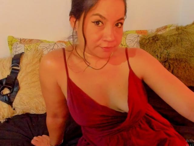 MsJustineBeaux with medium tits in a red dress, sitting on a bed, smiling at the camera, dark hair in a messy bun, warm light, soft pillows in the background.