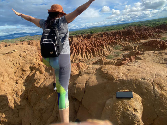 MelissaDelici se tient sur un terrain rocheux, les bras tendus, portant un chapeau marron, une chemise grise, des leggings colorés et un sac à dos noir, face à des formations rocheuses rouges, avec un téléphone sur le rocher en dessous..