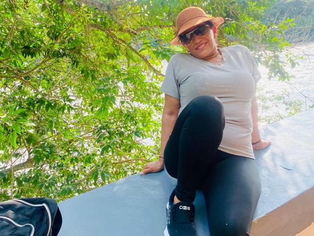 MelissaDelici with big tits, sitting on blue ledge, wearing gray top, black pants, brown hat, red sunglasses, and black C-3B sneakers, green leaves in background.