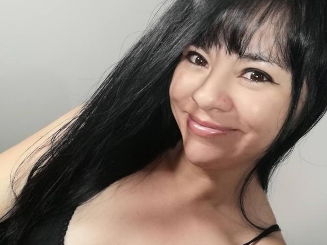 DayannaSquirt has long black hair, brown eyes, and a subtle smile with shiny pink lips. She's wearing a black top, showing her tits. Her skin is light.