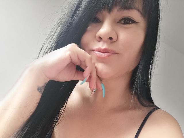 DayannaSquirt has long black hair, a nose ring, and blue acrylic nails, with a tattoo on her left hand, posing with her chin in her hand, and a slight smile.