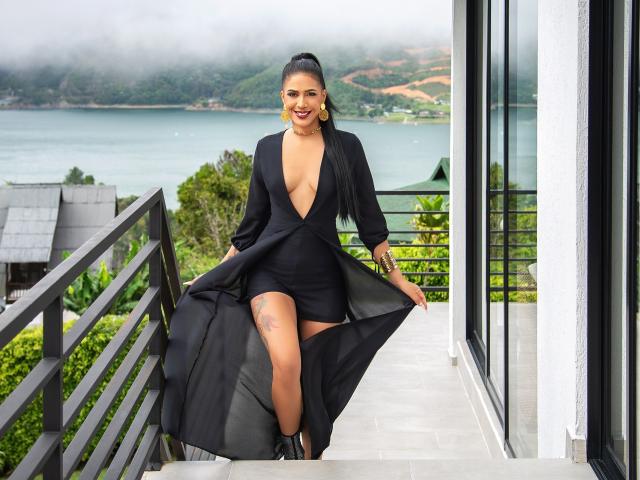 JoannaCool in a deep black dress with a massive ass, exposed tits, and a tattoo on her thigh, smiling, holding the dress, on a balcony with a misty view.