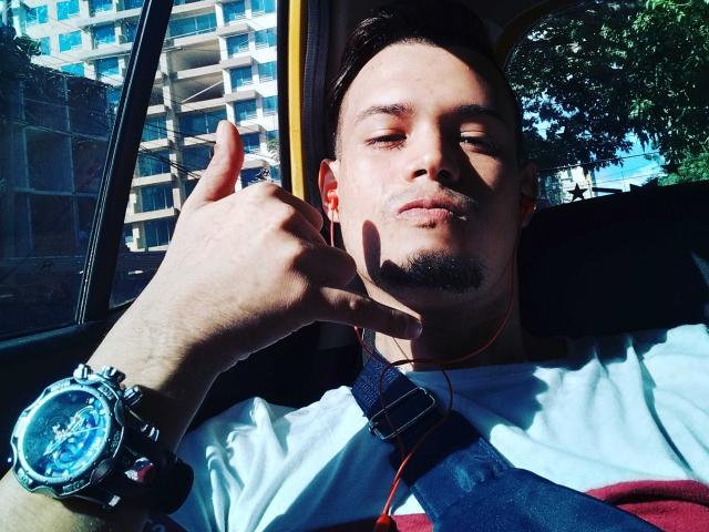 CarHot, con cabello corto y oscuro, piel clara y barba incipiente, levantando el pulgar, vistiendo una camisa blanca, una correa azul, auriculares naranjas y un reloj plateado con esfera azul..