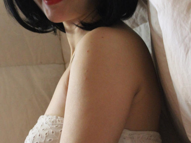 GloriaUpperLove's bare shoulder, with white lace top, showing her tits. She has red lipstick, black bob haircut, and light skin. The photo focuses on her upper body.