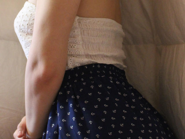 GloriaUpperLove's white, lace-strap top shows her small, perky tits. She's wearing a navy skirt with white anchor patterns, leaning forward, exposing her right arm.