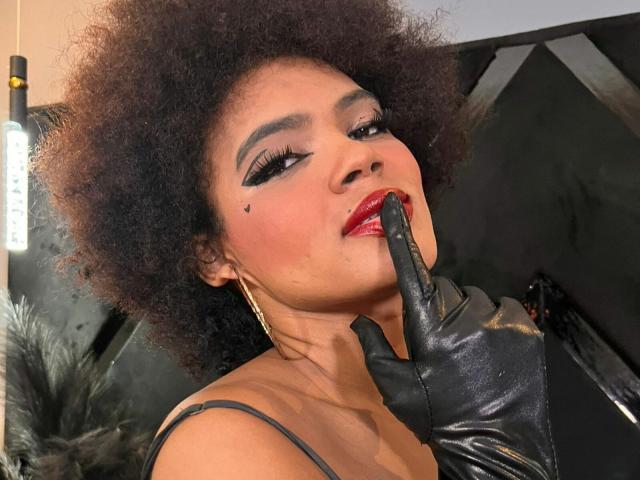 Alice Jelanni mit dunklen, lockigen Haaren, schwarzem Eyeliner, rotem Lippenstift, einem schwarzen Muttermal auf der Wange, einem schwarzen Handschuh, einem Finger in der Nähe der roten Lippen, goldenen Creolen, lächelnd, nach oben schauend..