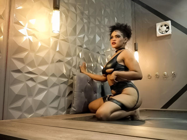 AliceJelanni, arrodillada, vestida con cuero negro, mostrando su trasero y sus pechos, con una correa en su muslo, frente a una pared geométrica, bajo una luz cálida, con una silla gris..