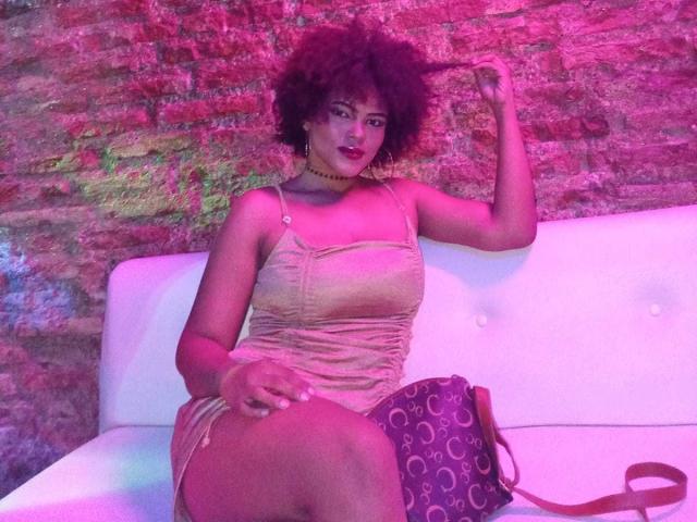AliceJelanni with dark curly hair, wearing a pink velvet dress, sitting on a white couch, holding her hair, purple bag on her right, pink and purple lighting.