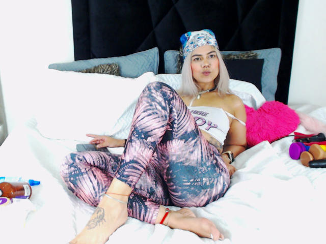 TarahDustin with blonde hair, wearing a blue headband, white crop top, and pink-black tie-dye leggings, sitting on a bed with a pink heart pillow, showing her ass.