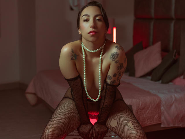KeitlyBlue, com tatuagens, vestindo meia-calça de rede preta, colar de pérolas e luvas, está sentada em uma cama, com as pernas afastadas, mostrando sua vulva, com uma luz vermelha atrás, olhando sedutivamente para a câmera..