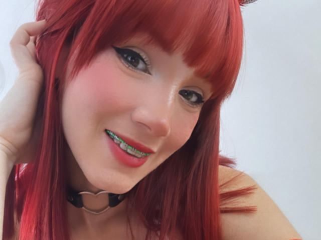 Akemi Haishi tiene el pelo rojo, delineador de ojos negro, brackets verdes, colorete rosa y un collar negro con un corazón. Está sonriendo y tiene una mano en el pelo..