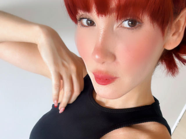 Akemi Haishi, con cabello rojo, labios rosados y piel clara, vestida con una camiseta negra sin mangas, con las manos cerca del pecho, mirando a la cámara con una ligera sonrisa..