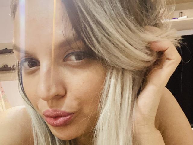 DouceAmelia's pouty face with pink lips, blonde hair, and brown eyes, holding her hair up, showing her nose and slight tan skin. Her hand's in her hair, looking cute.