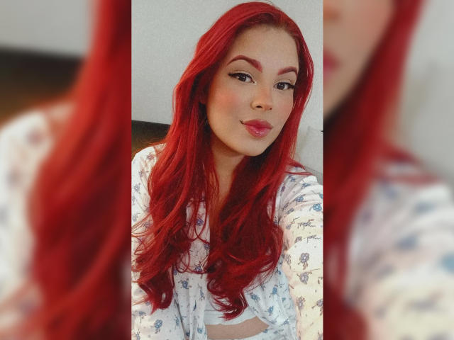 Selena Tyler, com cabelo ruivo, vestindo uma blusa branca com estampas florais, tem os seios levemente visíveis, lábios finos e um rosto bonito, refletida em um espelho, com reflexo borrado..