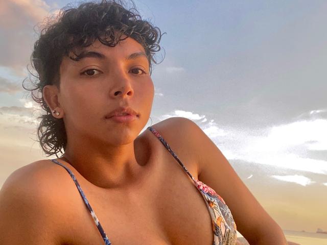 XHenisa a les cheveux noirs et bouclés, une peau brune, de petites boucles d'oreilles, et elle porte un haut de bikini à motifs floraux avec de fines bretelles, tout en regardant vers le bas la caméra..
