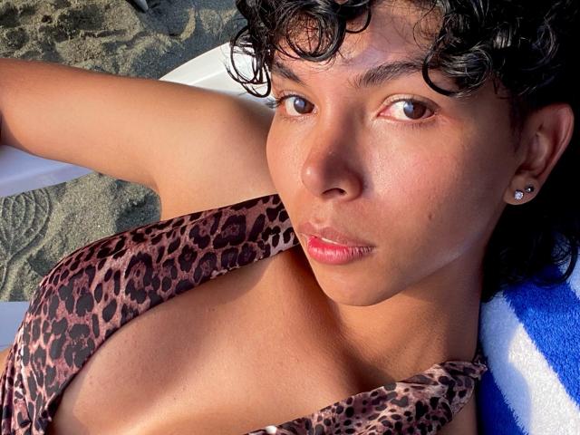 XHenisa, con i capelli neri ricci, gli occhi marroni e la pelle marrone media, indossa un bikini a stampa leopardo, è sdraiata su un asciugamano blu e bianco, mostrando il seno e le labbra rosa..