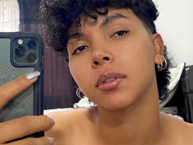 XHenisa, con i capelli neri ricci e corti, orecchini a cerchio e pelle marrone chiaro, tiene in mano un telefono, mostrando labbra leggermente divisa di colore rosato e un'espressione rilassata. Il seno non è visibile..