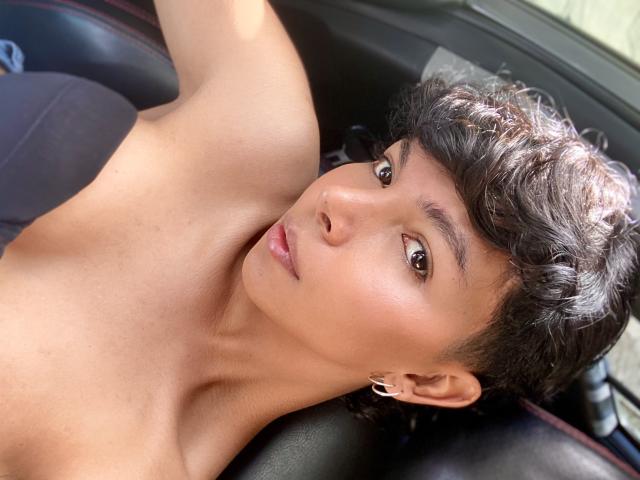 XHenisa tem cabelo preto encaracolado e curto, olhos castanhos e uma pele morena lisa. Ela está sem blusa, com um braço levantado, usando pequenas argolas nos ouvidos, e está deitada em um carro..