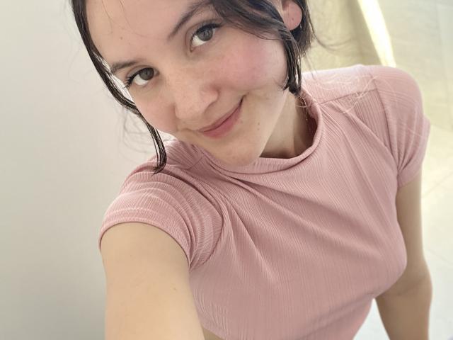 ChloeDulce, de piel clara, cabello castaño oscuro y pequeñas pecas, vestida con una camiseta rosa texturizada, sonriendo a la cámara, mostrando sus pequeños senos y su linda cara..
