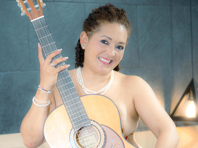FreyjaCam with curly brown hair, smiling, holding a guitar, wearing a pearl necklace and bracelets, topless, showing her tits, blue eyes, and a red lipstick.