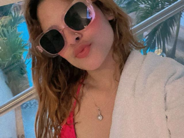 Shantal, com óculos de sol com tons de rosa, piercing no nariz e top de biquíni rosa, lábios curvados para baixo, usando uma toalha branca e um colar de prata, tem cabelo castanho ondulado e pele clara..