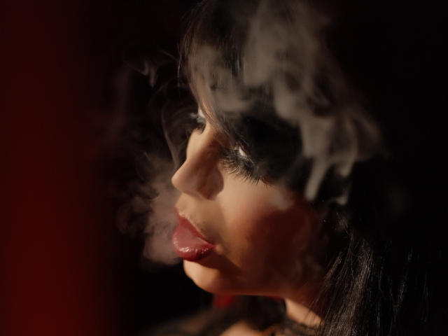 GabrielaAroms has smoke swirling around her face, with her tongue slightly out, dark lipstick, and black hair. Her nose is prominent, and her eyes are half-closed.