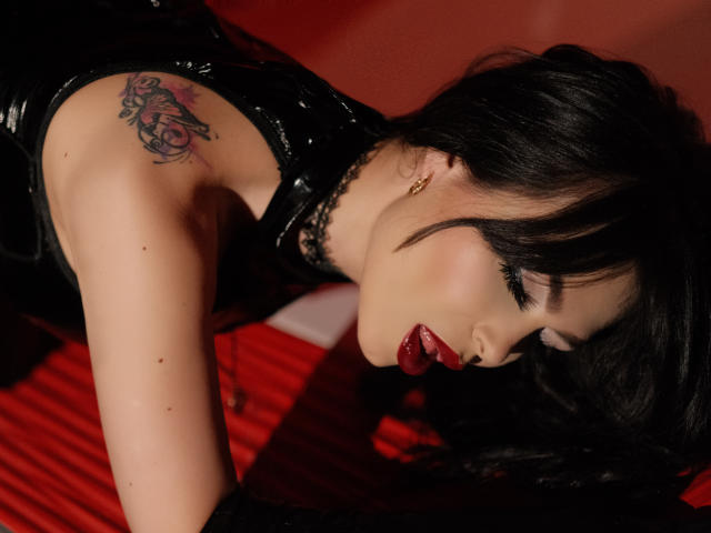 GabrielaAroms is on her knees, black hair down, red lipstick, black choker, tattoo on her shoulder, black latex, red background, eyes closed, sexy, and she's looking down.