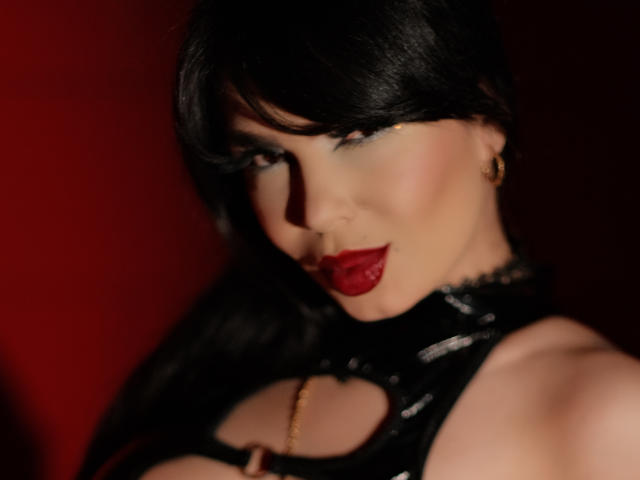 GabrielaAroms with dark hair, red lipstick, gold earrings, black cut-out top showing tits, and a seductive smile in a red-lit setting. Her eye makeup is subtle but sexy.